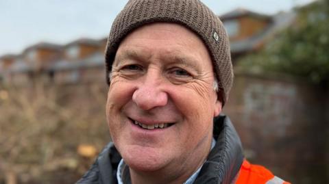 Andy Pratt smiles as he wears a high-vis jacket and brown woolly hat while standing outside.