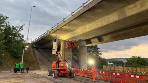Concrete bridge withe red and white barriers along the ground below it and several workers in high vis with a cherry picker vehicle.