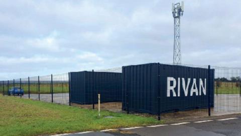 Two shipping containers are parked on a fenced in area on a field. One of the containers has white lettering spelling Rivan.