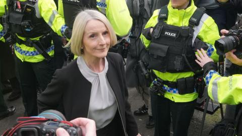 Lady Eleanor Donaldson, a woman with shoulder length blonde hair,  wearing a grey top and black jacket. She is surrounded by police officers and there are two cameras pointing at her. 