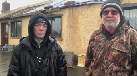 A woman and a man standing in front of a yellow bungalow. There is visible damage to the roof. They are wearing heavy rain jackets and hats.