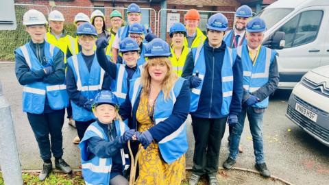 A group of people assembled outside a school while wearing hard hats and blue hi-vis jackets.