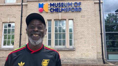A man wearing a Jamaica football shirt and a cap stands outside a building with a sign that says "Museum of Chelmsford". He is smiling at the camera.