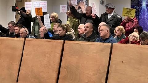 Three rows of people are sitting behind some wooden boards at Carlisle Civic Centre's main council chamber. Many are holding signs with messages such as "Cumbria Action says no to council of sanctuary"