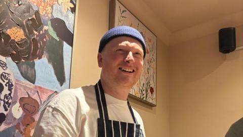 A man wearing a navy blue hat, a white t-shirt, and a navy blue apron with white stripes smiles with artwork behind him.