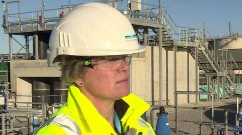 A woman is wearing a hard hat, goggles and a high-vis jacket and is standing by water treatment works