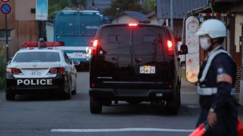 Police vehicles in a neighbourhood in the city of Nakano, where a murder suspect, believed to be a farmer in his 30s, was holed up inside a building. An officer can be seen in the foreground holding light batons.