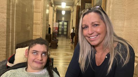 A blonde woman wearing a black top is smiling at the camera, beside her is her daughter Ellie who has dark hair and is sitting in a wheelchair