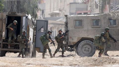 Five soldiers carry heavy weapons as they walk alongside two vehicles with rubble underneath their feet.