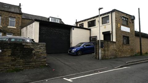 A garage which has a black roller shutter and a blue car parked on the drive outside. It is behind what looks like a row of stone terraced houses. 