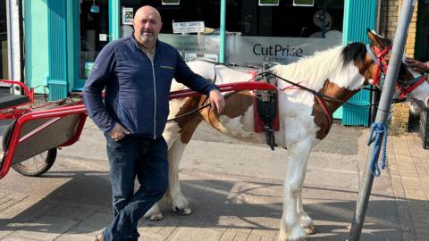 Man wears a blue jumper stands alongside a horse and cart outside a shop front. the shop sells carpets. The horse is white and brown. A hand is stroking the horse's forelock. The cart is an open one and is painted red. 