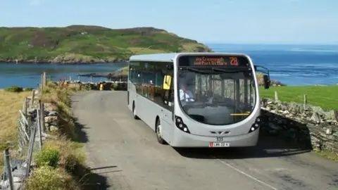 A modern silver bus on a country road with a headland on the Calf of Man and blue seas behind it. There are Manx stone walls and fencing running alongside green fields on either side of the road.