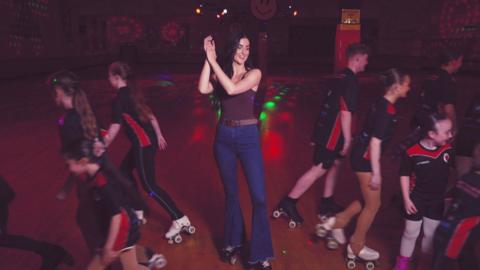 Presenter Vicky Hawkesworth claps in the middle of a roller rink, surrounded by kids setting off skating