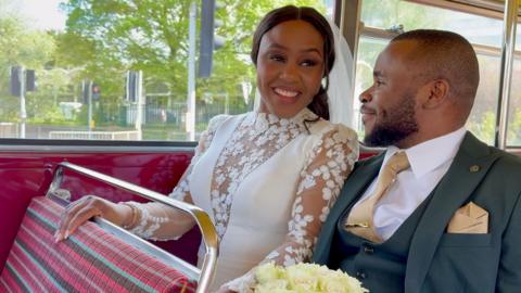 A woman wearing a wedding dress is smiling next to a man in a suit as they ride inside a 1963 double decker bus.