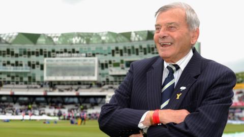 A man with grey hair stands with his arms folded at the edge of a cricket pitch. He is smiling and wearing a pin stripe navy suit, with a white shirt and striped nay tie. Behind him a large cricket stand can be seen in the distance.
