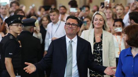 Andy Burnham, wearing a dark suit, silver tie and white, with arms open and a crowd behind him
