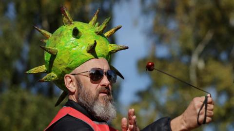 A bearded man wearing a horse chestnut shell model hat on his head. He is playing conkers with a conker on the end of a black string. 