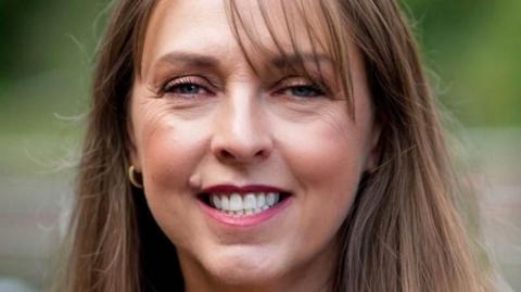 A woman with long brown hair, a side fringe, wearing a black leather jacket and white T-shirt smiling at the camera