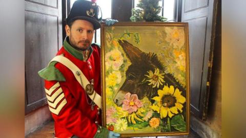 Dr Robert MacKinnon is in a red and green jacket with a white strap and medals pinned to it. He is holding a painting in a gold frame. The painting is of a brown donkey surrounded by flowers, on a yellow/green background.