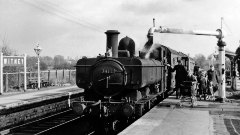 A black and white photo shows an old steam engine pulled up at Witney train station. Passengers are getting on, including a woman with a pram and a family with a young child.