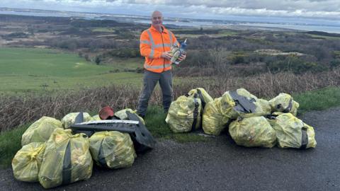 Frank Roberts stood at the side of the road surrounded by yellow rubbish bags. He is wearing an orange high visibility jacket and is stood in front of a green field.
