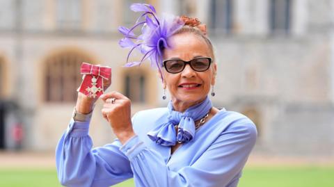 Anita Neil, who is wearing a purple blouse, scarf and fascinator, smiles as she holds up her award in the grounds of Windsor Castle