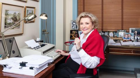 Barbara Taylor Bradford sat at a desk with a red jumper draped over her shoulders. She smiles at the camera and is holding a red pair of glasses in one hand as her other rests on a typewriter upon the desk in front of her. Behind her is another desk which has a display of framed photos.