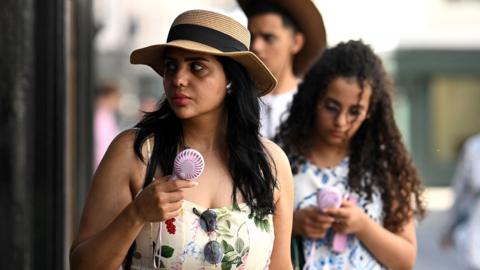 People walk down a street carrying portable electric fans and looking hot, while wearing summer clothing, in York on 24 June.