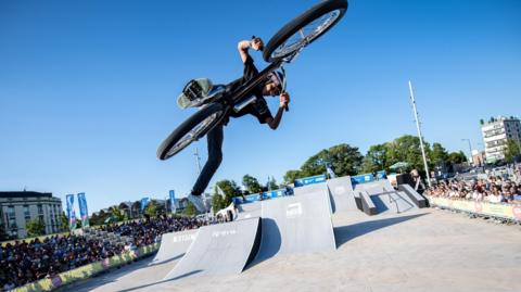 A BMX rider in mid-air, holding on to his bike with one foot trailing behind. In the background, there are BMX ramps and a crowd of onlookers. 