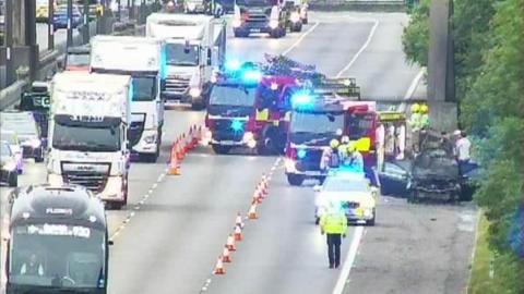 A motorway, with traffic cones on it and two fire engines, a burned out vehicle to the left, and cars travelling past a number of cones. Emergency workers are walking on the road. There are queues of traffic. 