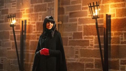 Picture of Claudia Winkleman in The Celebrity Traitors. She is in a black cloak and wearing red gloves. There arelanterns with flames behind her