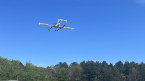 A drone comes in to land in a field with trees in the background. 