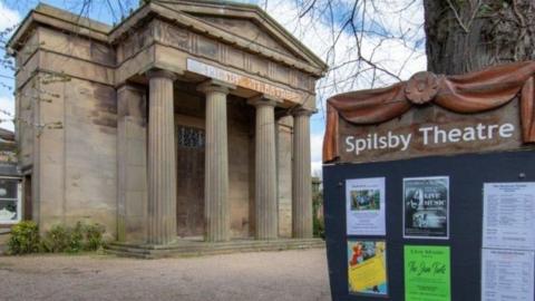 The exterior of Spilsby Sessions House. It is a historical building with four large columns at the front. A sign in the foreground reads "Spilsby Theatre" and has some posters on it with details of events. 