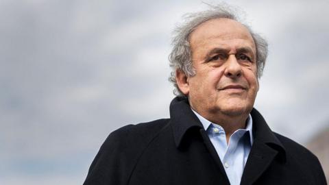 Michel Platini, silver-haired and wearing a black coat over a pale blue shirt, looks on