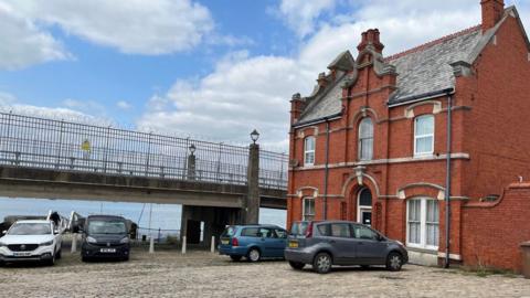 A red brick house on the rifht. There is a footbridge. There are four cars parked. The sea is a light blue. The sky is blue with clouds. 