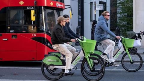 The image shows three people riding Lime-branded electric bikes along a busy city street. The bikes are the familiar green-and-white design used in Lime’s rental fleet. red double‑decker bus is behind them, suggesting a central urban area with mixed transport options. A street sign reading “Durham House Street WC2” places the scene in central London.