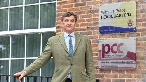 Philip Wilkinson in a white shirt, blue tie and green suit. He stands in front of a brick building which includes a sign for Wiltshire Police and the Police and Crime Commissioner. 