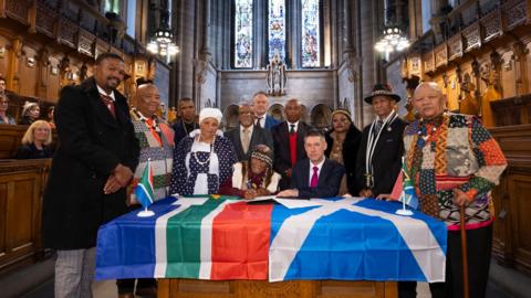 Several people in a chapel, surrounding a table at which two people are seated. The South Africa and Scottish flags are draped over the table. One of the men is using a pen to sign something, while the others - numbering about 10 watch on. Other people watch from the chapel's pews.