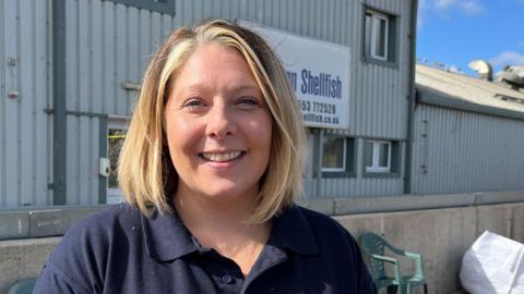 Abbie Williamson standing outside with a large grey building behind her. She has shoulder-length blonde hair, is wearing a blue polo-shirt and smiling. 