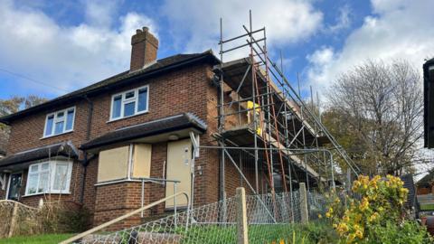 A semi-detached terraced house with boarded up ground floor windows and scaffolding on its right wall