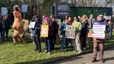 A group of people holding cardboard signs at a picket line with one person dressed like an orange dinosaur in an inflatable costume. They are standing in front of the blue Heriot Watt University Edinburgh campus sign