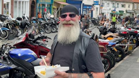 A person with a long white beard stands among rows of parked motorcycles at a busy biker gathering, holding a tray of chips in a bustling town street.