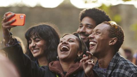 A woman holds up a mobile phone in an orange case. She and her friends laugh and smiled for a selfie