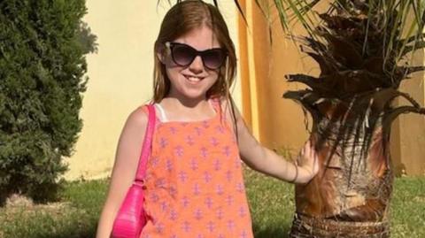 Edie Smart is smiling on a sunny day leaning against what looks like a palm tree. She is wearing an orange, sleeveless top and has a pink bag over her shoulder. She is wearing big sunglasses and has fair, long hair.
