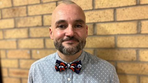 A bald man wearing a grey shirt with a white pattern and a red black and white bow tie, smiles as he stands in front of a brick wall.