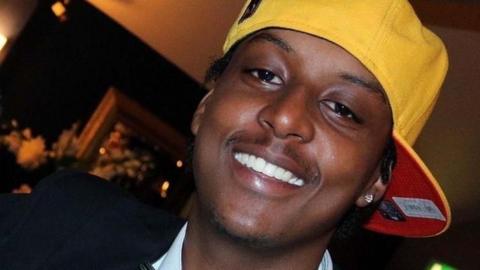 A close up image of Eddie Kinuthia smiling at the camera. He is wearing a white collared shirt, a black blazer and a yellow cap with a red underside. His hair is twisted into short locks. 