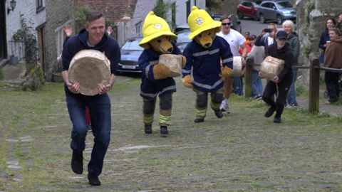 Two men flanking two people dressed in teddy bear costumes, who are dressed as firefighters, running up the hill. 