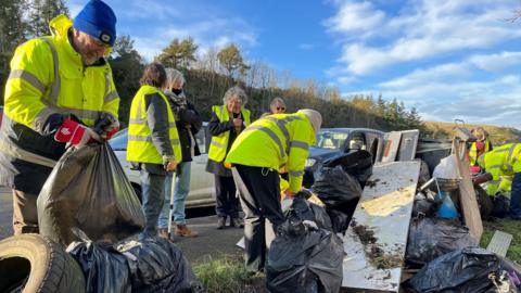 A group of eight people in hi-vis jackets tying up black waste bags with vehicles in the background