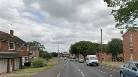 A long street heading away from the camera. There are cars and a bus on the right hand side of the road. There are houses and green grass on either side