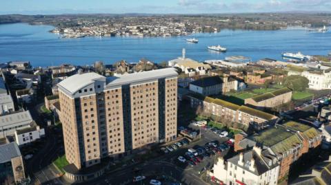 A drone image of the existing building which is set to be replaced. It is a large brown high rise 14-storey building with grey roof. It towers over the buildings set around it. A car park wraps around the building.Plymouth Sound is a short distance away and buildings can be seen on the opposite back. There are three ferries crossing the water. 